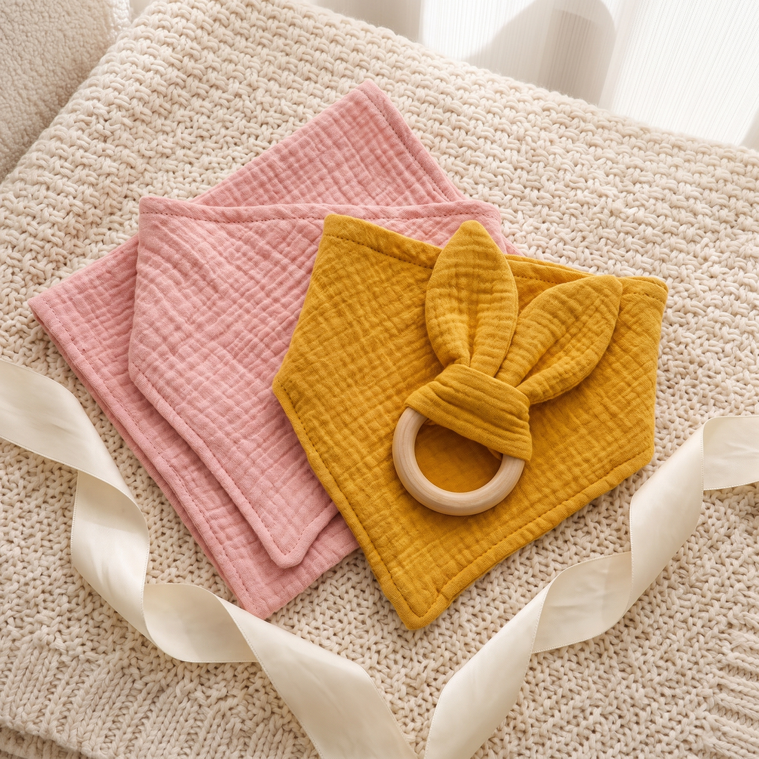 Muslin bib, square cloth and bunny-ear wooden teething ring, arranged as a gift set on a cream blanket